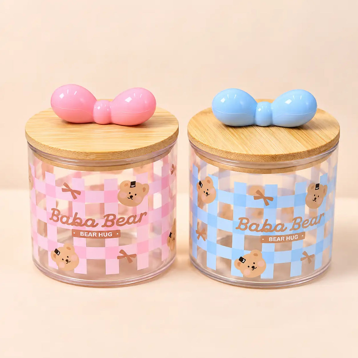 Two cute cylindrical storage jars with bamboo lids, decorated with pink and blue checkered patterns and cartoon bear designs. Each jar has a wooden bow-shaped lid topper, one pink and one blue.