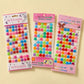 Three packages of colorful craft embellishments labeled 'Say it in cry stasis' on a light beige background. The left package contains pastel and bright star-shaped crystals, the middle contains round multicolor beads, and the right package contains round multicolor stickers.