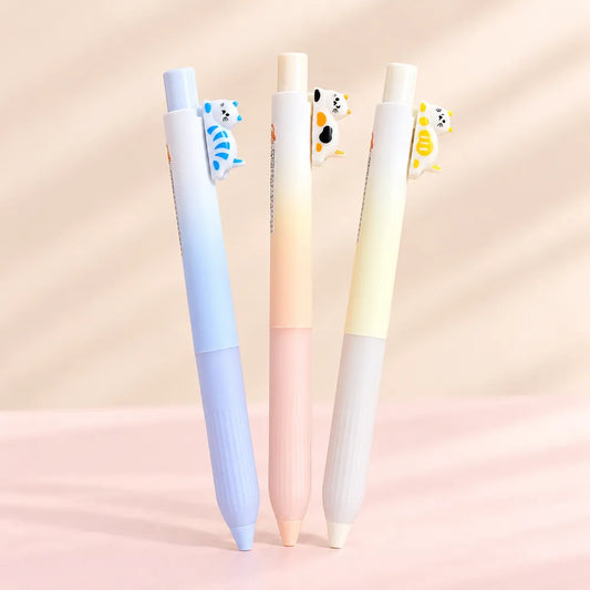 Three pastel-colored gel pens standing upright on a soft pink surface against a warm beige background. Each pen has a small cartoon animal clip attached near the top.