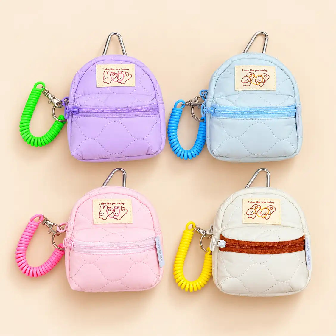 Four quilted mini backpack keychains arranged in a 2x2 grid on a light beige background. Each backpack has a different pastel color: lavender purple, light blue, pink, and off-white.
