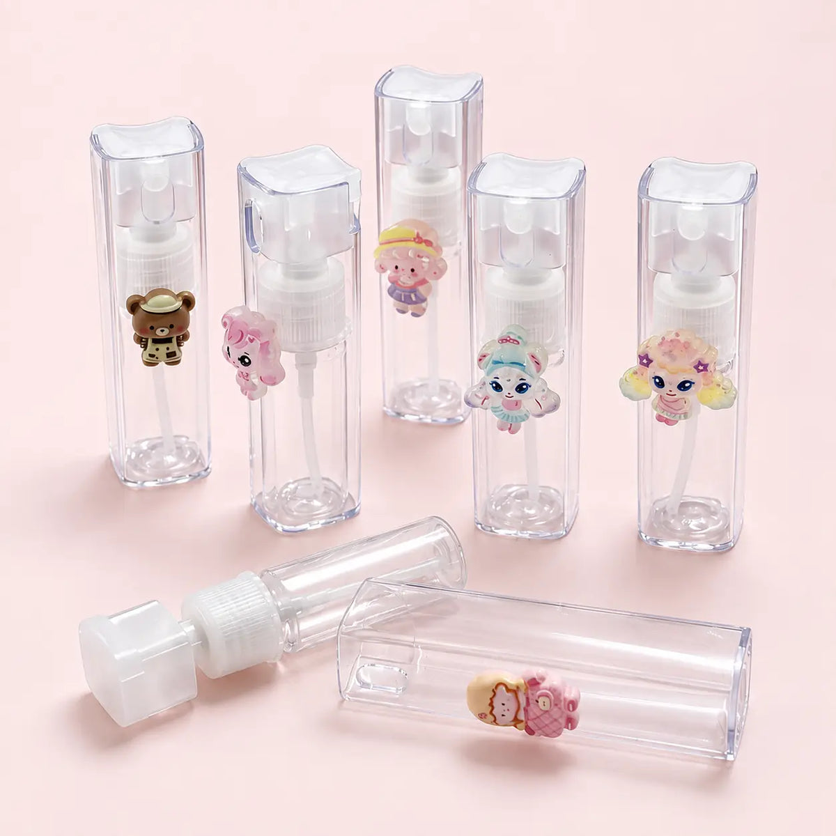Several small clear plastic spray bottles with white pump caps are arranged on a pale pink background. Each bottle has a small colorful cartoon character attached near the top, and one bottle is shown uncapped with its cap placed beside it.