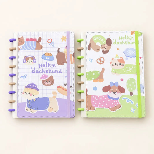 Two spiral-bound planner notebooks with cute dachshund illustrations, shown side by side on a light cream background. The left notebook has a lavender grid cover with purple spiral rings, and the right notebook has a green cover with green spiral rings.